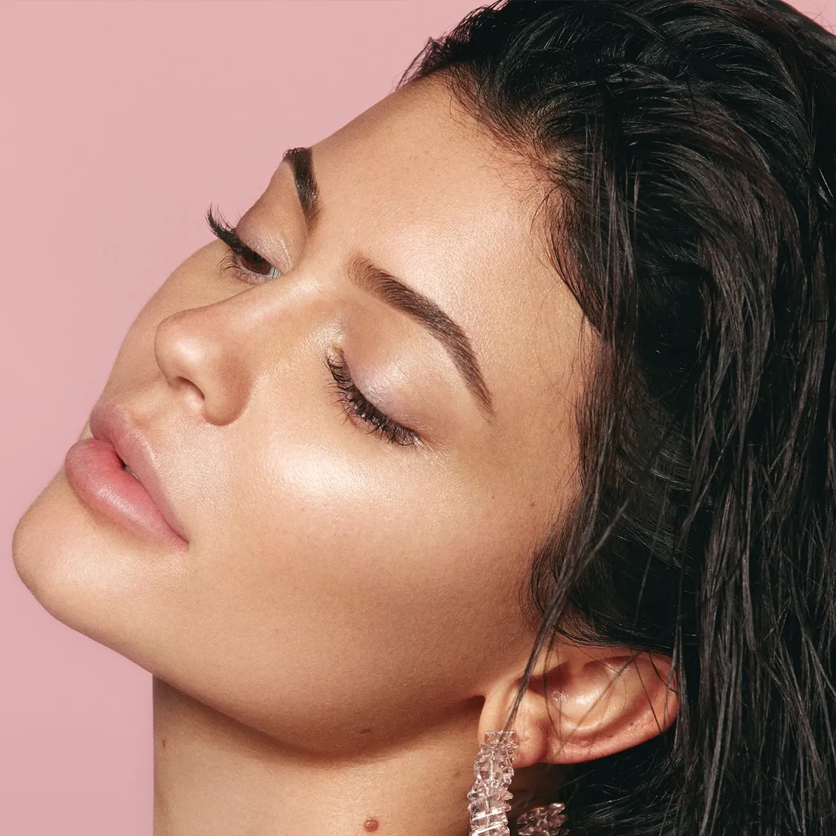 Kylie Jenner i profil som ser ned og lener hodet bakover mot en rosa bakgrunn.