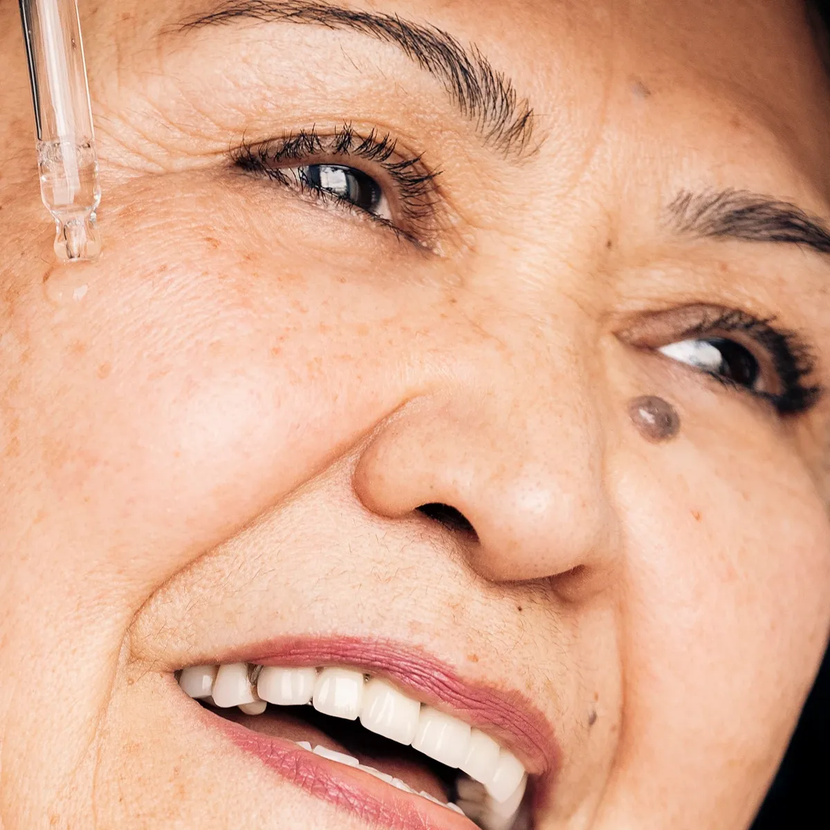 Nærbilde av en smilende eldre kvinne som påfører serum under øyet med en pipette.