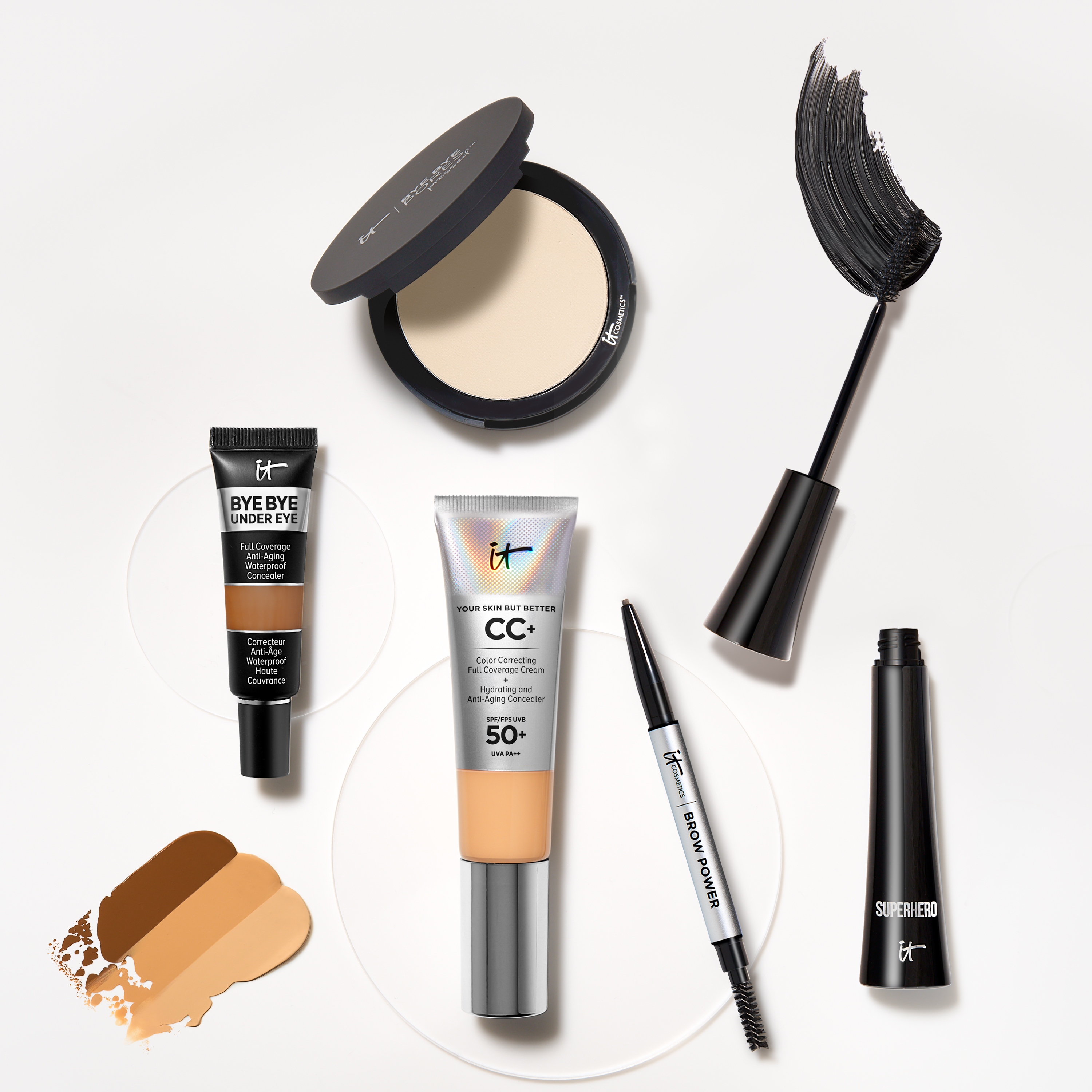 Flera sminkprodukter från IT Cosmetics. Produkterna är arrangerade på en ljus yta med färgprover av concealer och CC+ kräm.