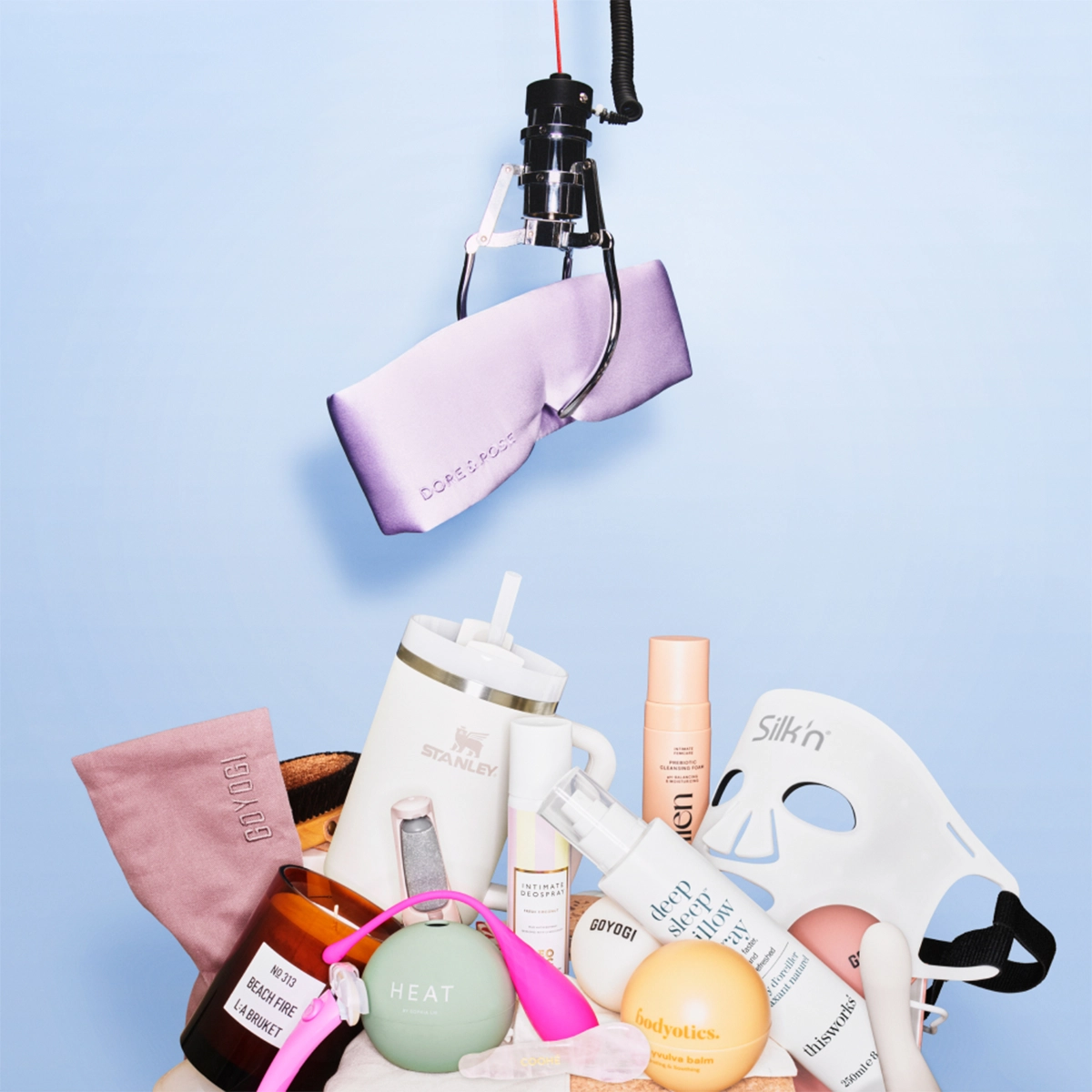 Skjønnhets- og wellnessprodukter arrangert mot lys blå bakgrunn med en robotarm som holder en lilla DORE & ROSE-satinmaske. Bildet viser blant annet hudpleie, sexleketøy, body lotion, Silk’n LED-maske, parfyme fra BYREDO og duftlys fra La Bruket.