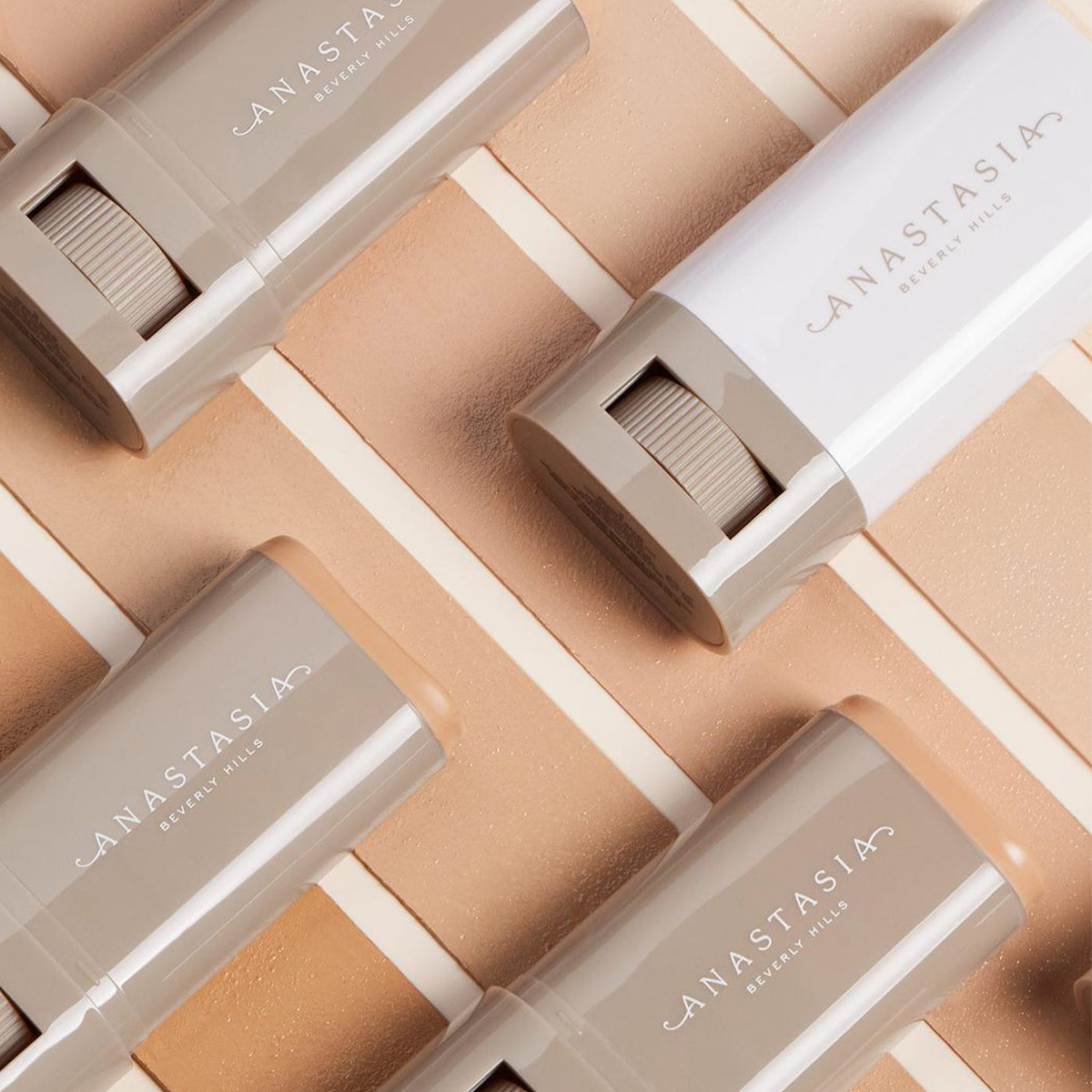Foundationstift från märket Anastasia Beverly Hills. Stiften är placerade diagonalt över en bakgrund med olika nyanser av beige och brunt, vilket representerar olika hudtoner. Varje stift har en grå och vit förpackning med märkesnamnet tryckt på sidan. Bakgrunden har en texturerad yta som framhäver produkterna och deras färger.