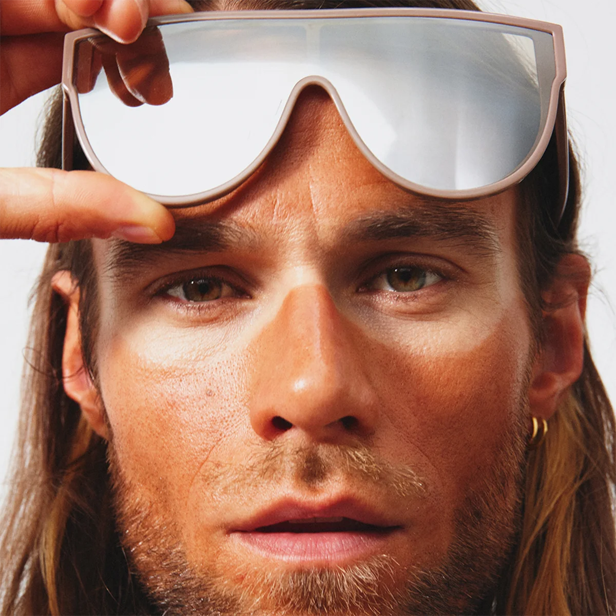 En person løfter et par store, speilblanke solbriller over øynene, noe som fremhever tydelige solbeskyttelseslinjer og kontrast mellom solbrune og beskyttede partier i ansiktet, med fokus på hudpleie og SPF.