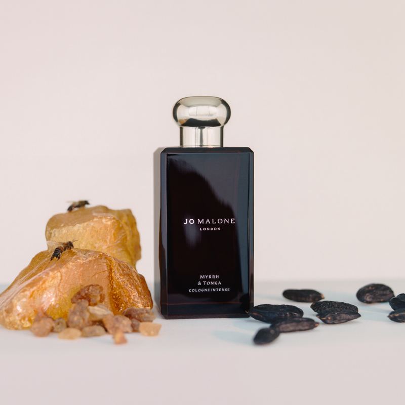 En svart parfymeflaske fra Jo Malone med teksten «Myrrh & Tonka Cologne Intense» på hvit bakgrunn. Ved siden av flasken er det myrrasteiner og tonkabønner.