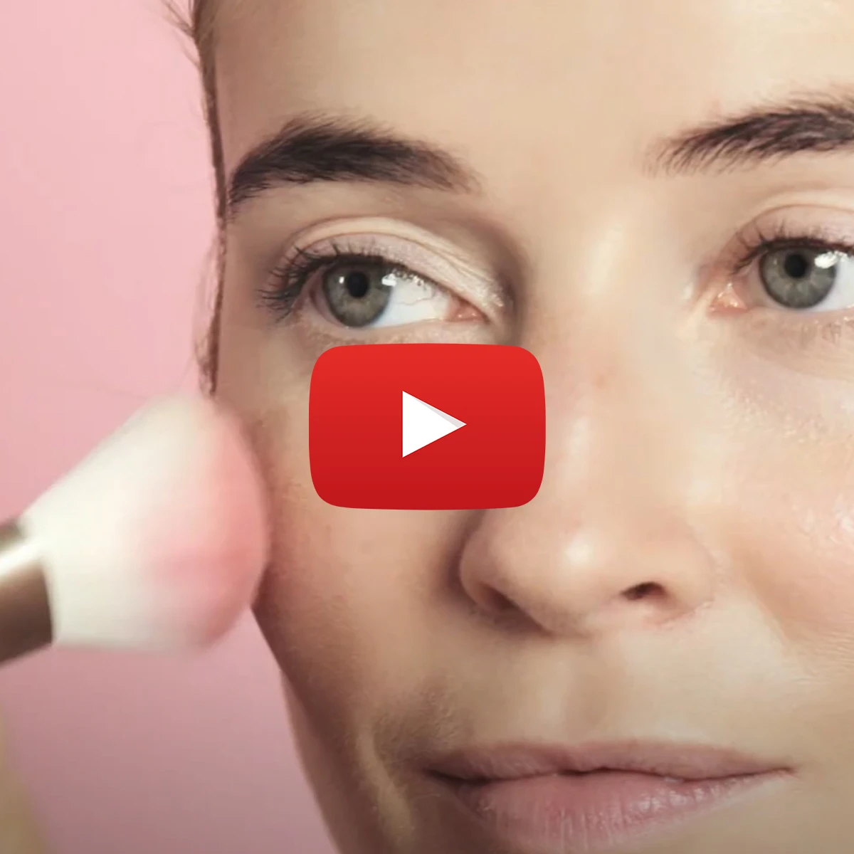 Nærbilde av en kvinne som påfører blush med en sminkebørste på kinnet. En rød YouTube-avspillingsknapp vises over bildet