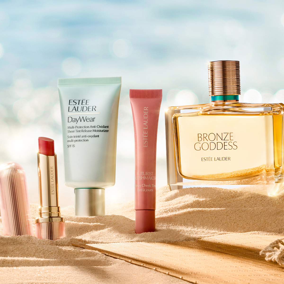 Neljä Estée Lauder -tuotetta on aseteltu hiekkaan vaalean ranta- ja merimaisemaa muistuttavan taustan eteen. Kuvassa näkyvät Estée Lauder -huulipuna, DayWear Multi-Protection Anti-Oxidant Sheer Tint Release Moisturizer SPF 15, Futurist Soft Touch Brightening Skincealer sekä Bronze Goddess -hajuvesi, jossa on näkyvä teksti ”BRONZE GODDESS ESTÉE LAUDER”.
