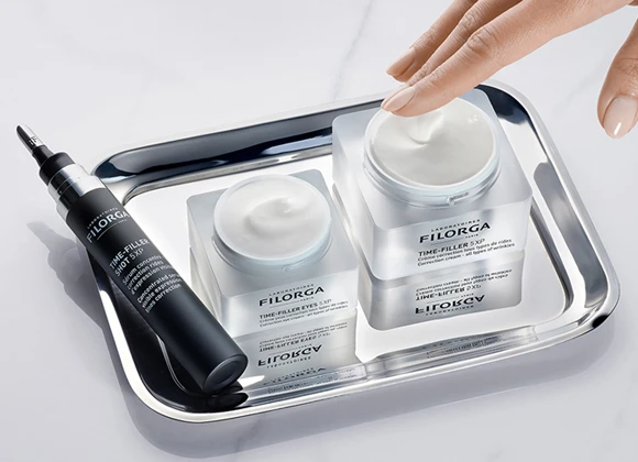 En hånd berører en krukke med Filorga "TIME-FILLER 5XP" krem, som står på et sølvbrett sammen med en mindre krukke og en tube med "TIME-FILLER EYES 5XP".