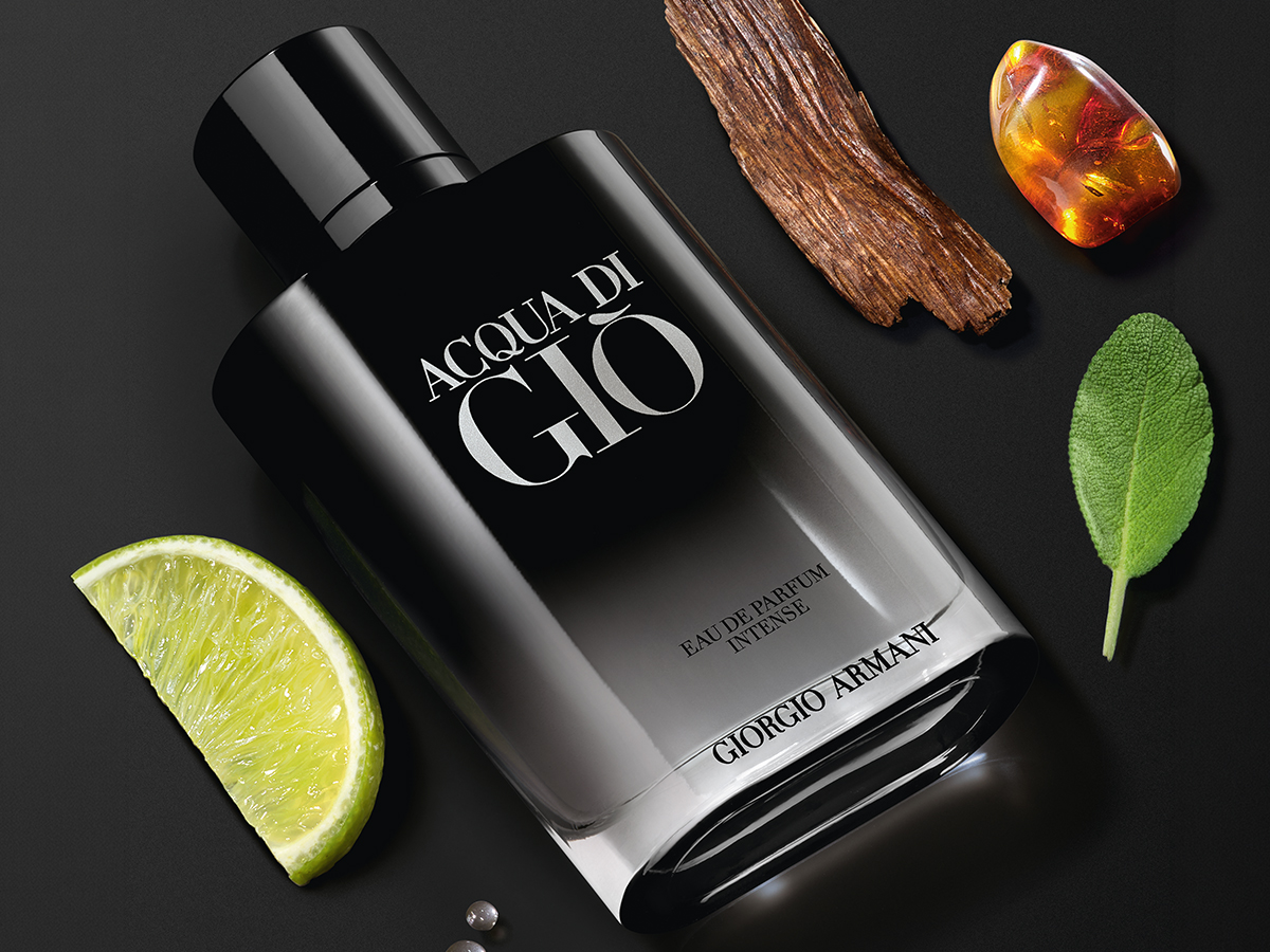 Sort Acqua di Giò-parfymeflaske (Giorgio Armani Eau de Parfum Intense) på mørk bakgrunn, omgitt av en limebåt, et stykke tre, en ravfarget stein og et grønt blad.