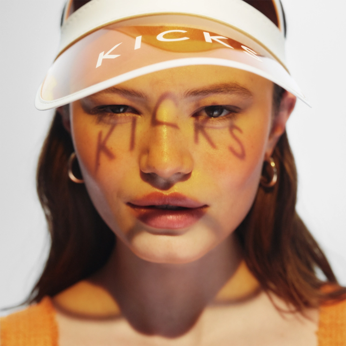 Närbild av ansikte med kreativ makeup i gula och orange toner samt glansiga läppar, där ordet “KICKS” är målat över kinder och näsa. Personen bär en transparent solskärm med texten “KICKS” och stora hoop-örhängen.