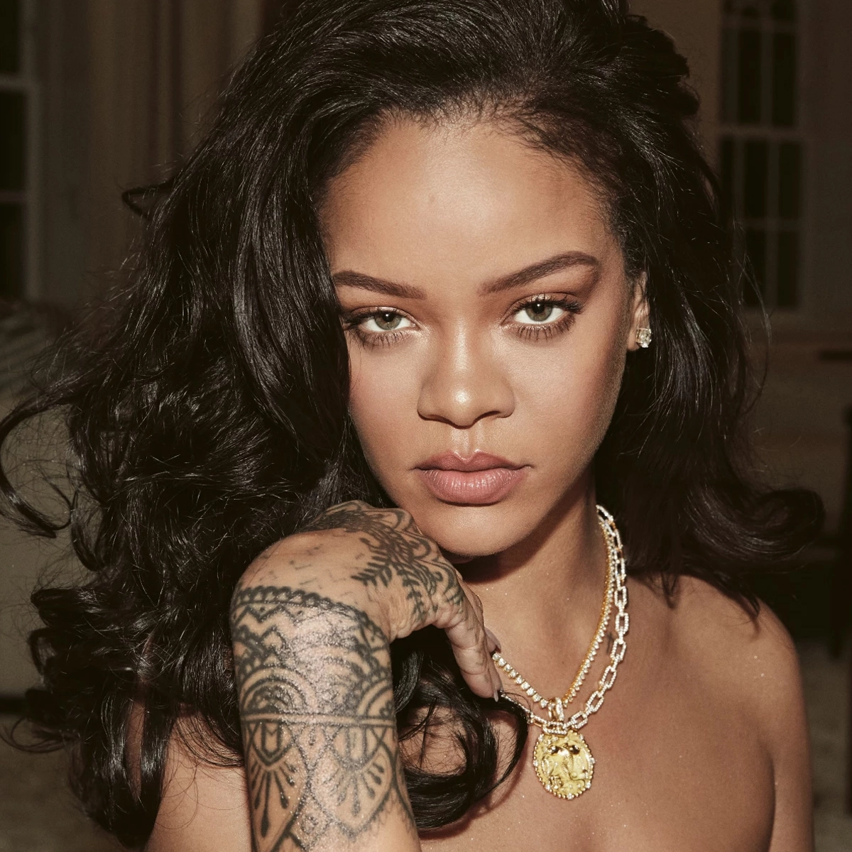 Rihanna tittar in i kameran iklädd halsband och visar hand med tatueringar. Sitter i vardagsrumsmiljö.