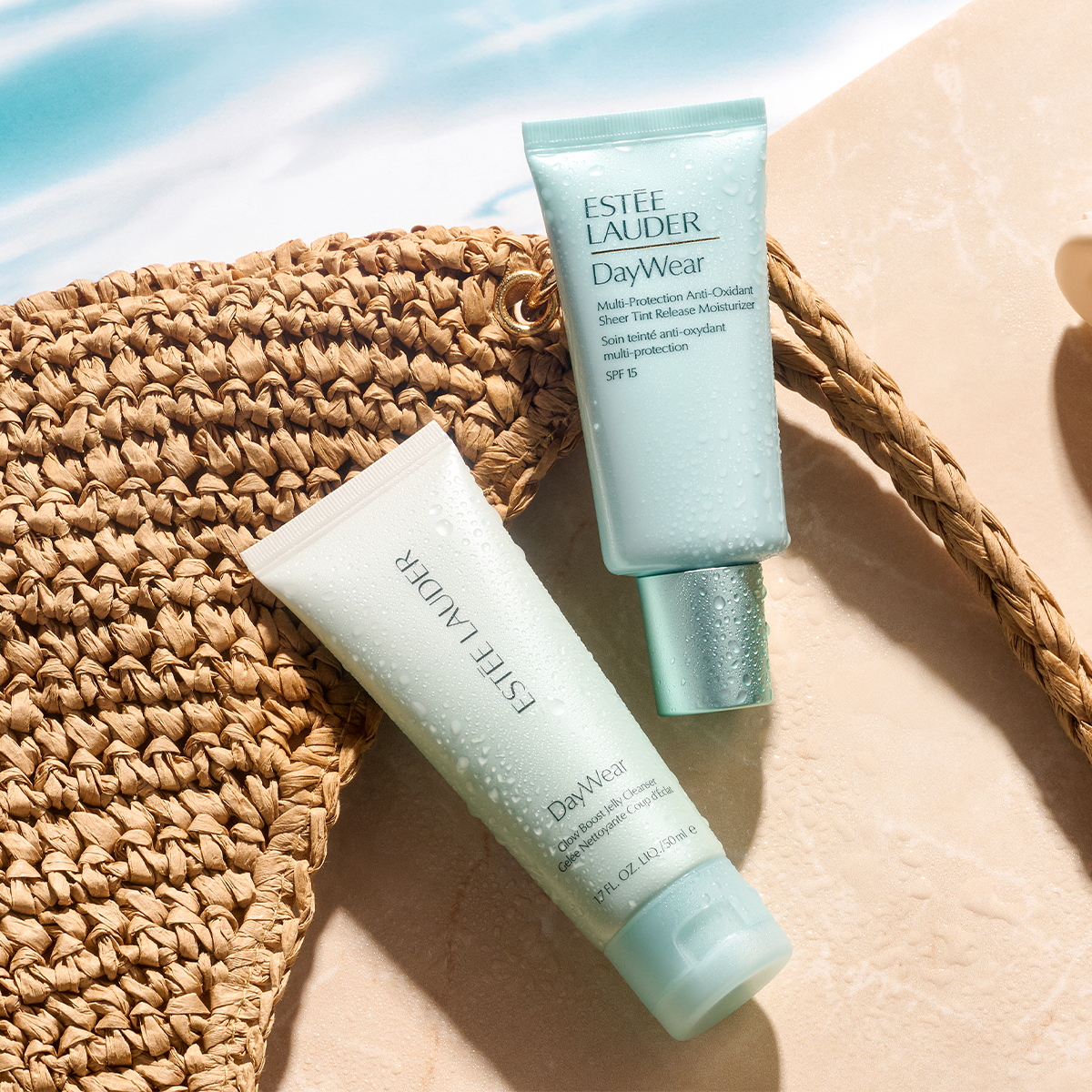 To Estée Lauder DayWear-hudpleieprodukter ligger på en sandfarget overflate ved siden av en flettet strandveske. Tubene er dekket med vanndråper og viser tekstene «ESTÉE LAUDER DayWear Multi-Protection Anti-Oxidant Sheer Tint Release Moisturizer SPF 15» og «ESTÉE LAUDER DayWear Glow Boosting Jelly Cleanser 150 ml».