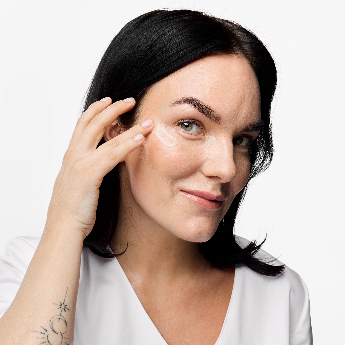 Person påfører øyekrem eller hudpleiekrem under øyet med fingertuppene. Nærbilde med naturlig makeup, mørkt hår og hvit topp mot lys bakgrunn.