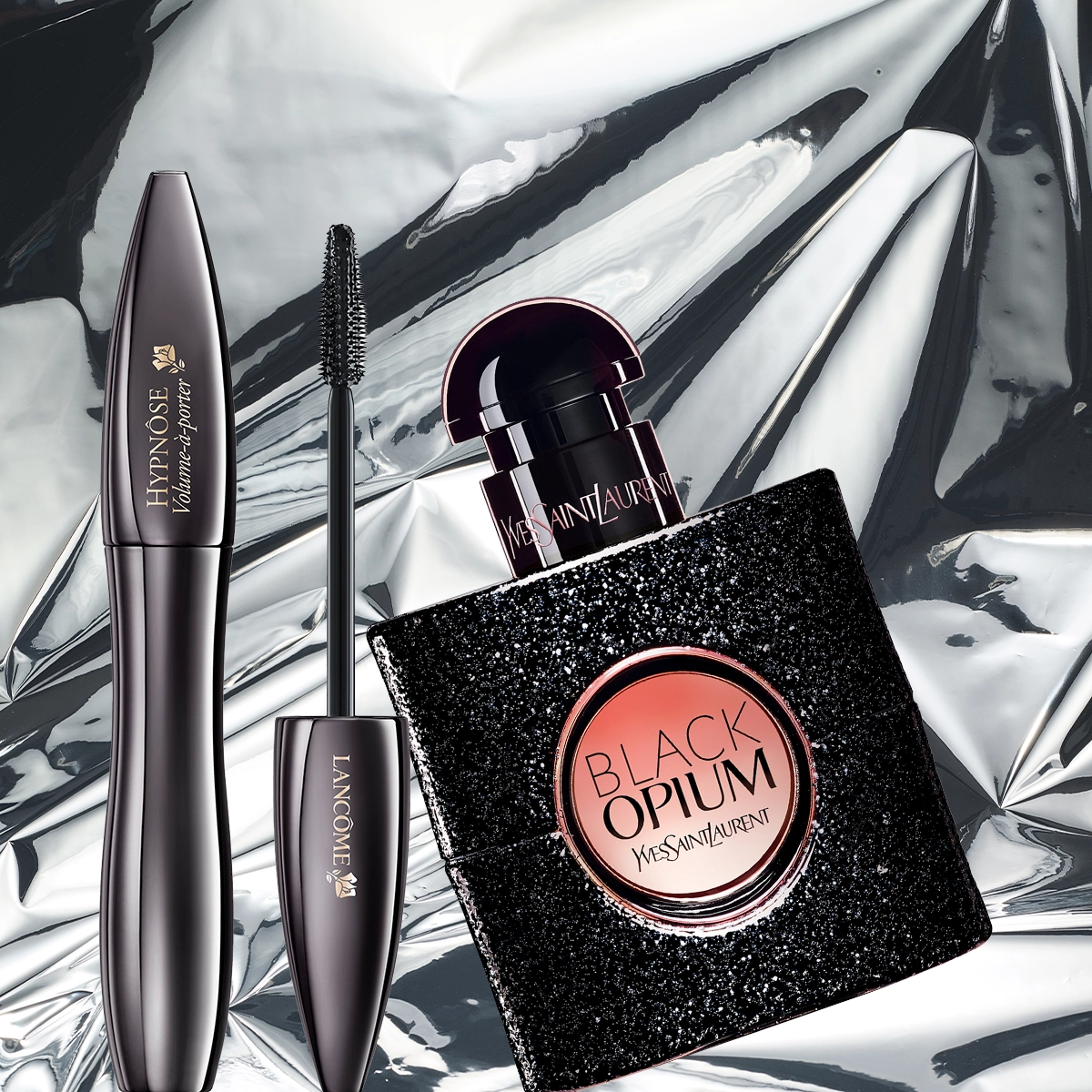 Lancôme Hypnôse Volume-maskara og Yves Saint Laurent Black Opium-parfyme er plassert mot en blank sølvbakgrunn som fremhever en glamorøs beautystemning.
