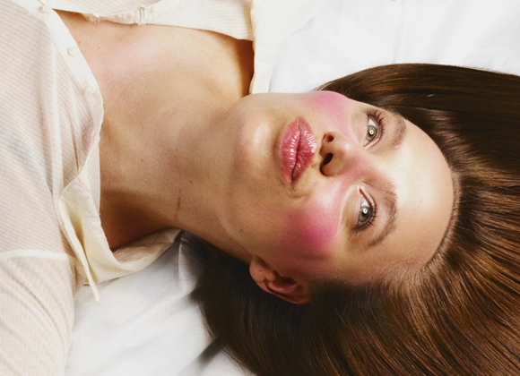 En person ligger på ryggen på hvitt sengetøy med langt, brunt hår, rosa kinn og en lys skjorte.