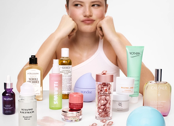Person som hviler haken i hendene bak et bord fylt med hudpleie- og skjønnhetsprodukter, inkludert L’Occitane Néroli & Orchidée, Kiehl’s Calendula Herbal Extract Toner, Biotherm Aquasource, NUXE Huile Prodigieuse, Caudalie Eau des Vignes, Medik8 serum, Susanne Kaufmann Deodorant Roll-On og Elizabeth Arden kapselserum.