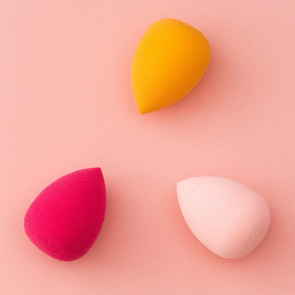 Tre sminksvamper i forskjellige farger (gul, rosa og rød) på en rosa bakgrunn.