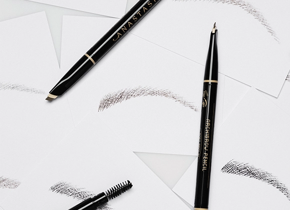 Svart ögonbrynspenna från Anastasia Beverly Hills med texten "ANASTASIA" och "BROW WIZ", tillsammans med en spoolieborste och flera streck av brynfärg på vit bakgrund.