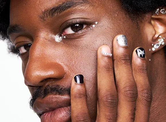 Lähikuvassa näkyy henkilö tummalla ihonsävyllä ja luonnollisilla kiharoilla hiuksilla, kasvot osittain kättä vasten. Kynnet ovat koristeltu mustalla lakalla, hopeisella glitterillä ja taiteellisilla yksityiskohdilla, kuten tähtikuviolla ja pienillä kristalleilla, jotka tuovat ilmeeseen ylellistä ja trendikästä tunnelmaa. Silmien ympärillä näkyy hohtavaa meikkiä sekä poskipäälle aseteltuja pieniä kimaltelevia koristeita, jotka luovat rohkean ja juhlavan lookin. Kuvan kokonaisuus huokuu modernia kauneutta, luovaa itseilmaisua ja näyttävää kynsityyliä.