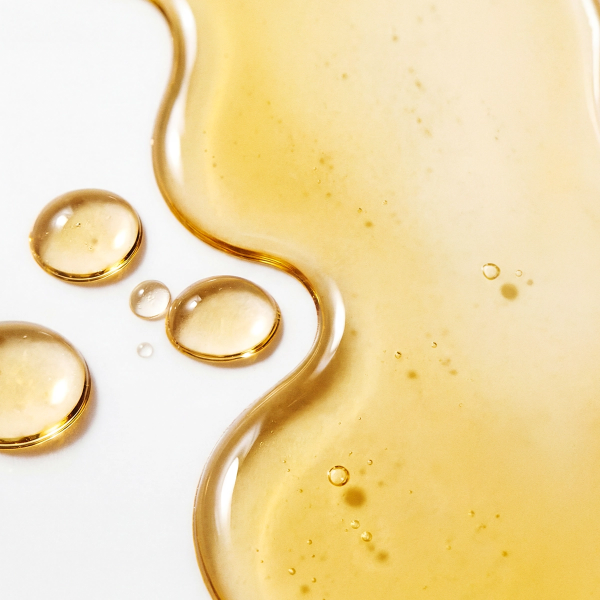 Nærbilde av gyllen ansiktsolje eller serum i flytende form med dråper og myke former på en lys bakgrunn, med fokus på hudpleie og fuktighetsgivende serum.