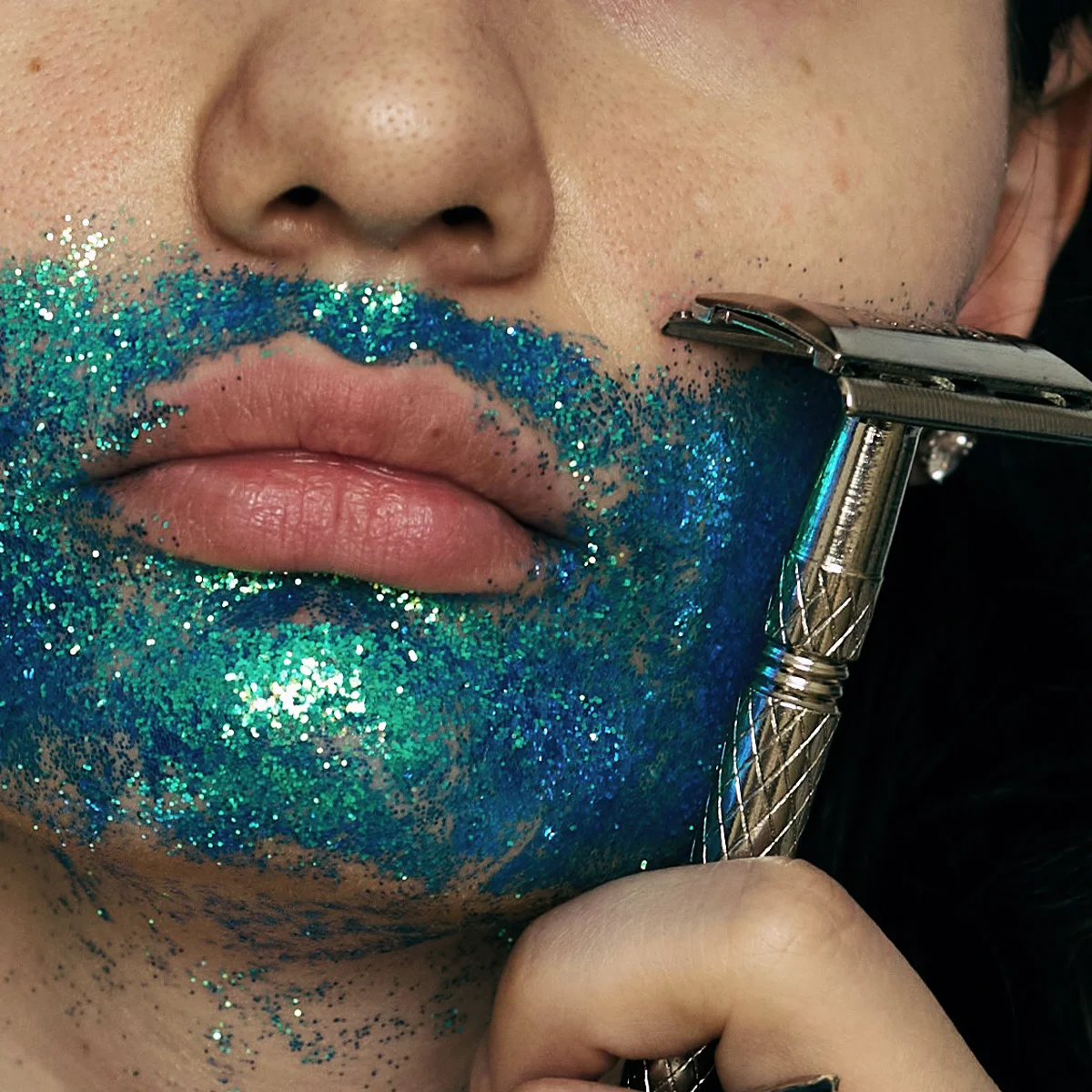 Nærbilde av munn og hake med blågrønt glitter på huden, der en metallbarberhøvel holdes mot haken under barbering mot mørk bakgrunn.