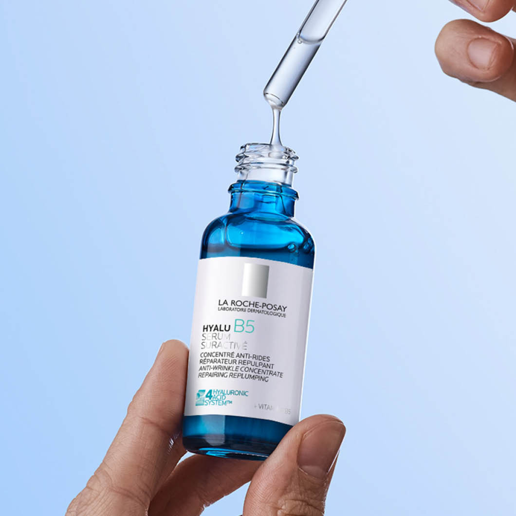En hand håller en blå glasflaska med pipett från La Roche-Posay medan serum droppas ner i flaskan. Texten på flaskan visar “LA ROCHE-POSAY HYALU B5 SERUM”, “ANTI-WRINKLE CONCENTRATE REPAIRING REPLUMPING” och “HYALURONIC ACID”.