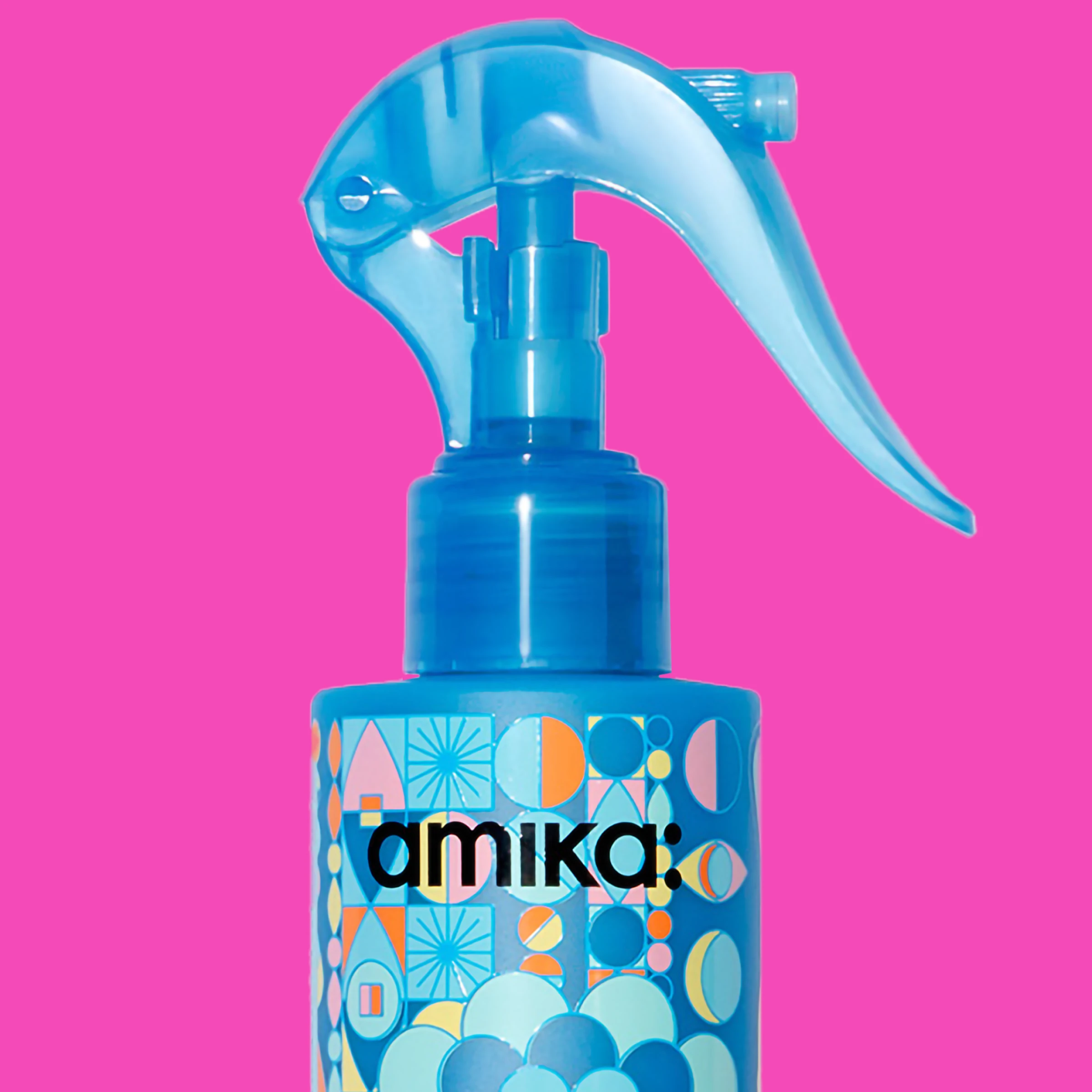 En blå sprayflaske fra Amika med fargerike geometriske mønstre mot en knallrosa bakgrunn.