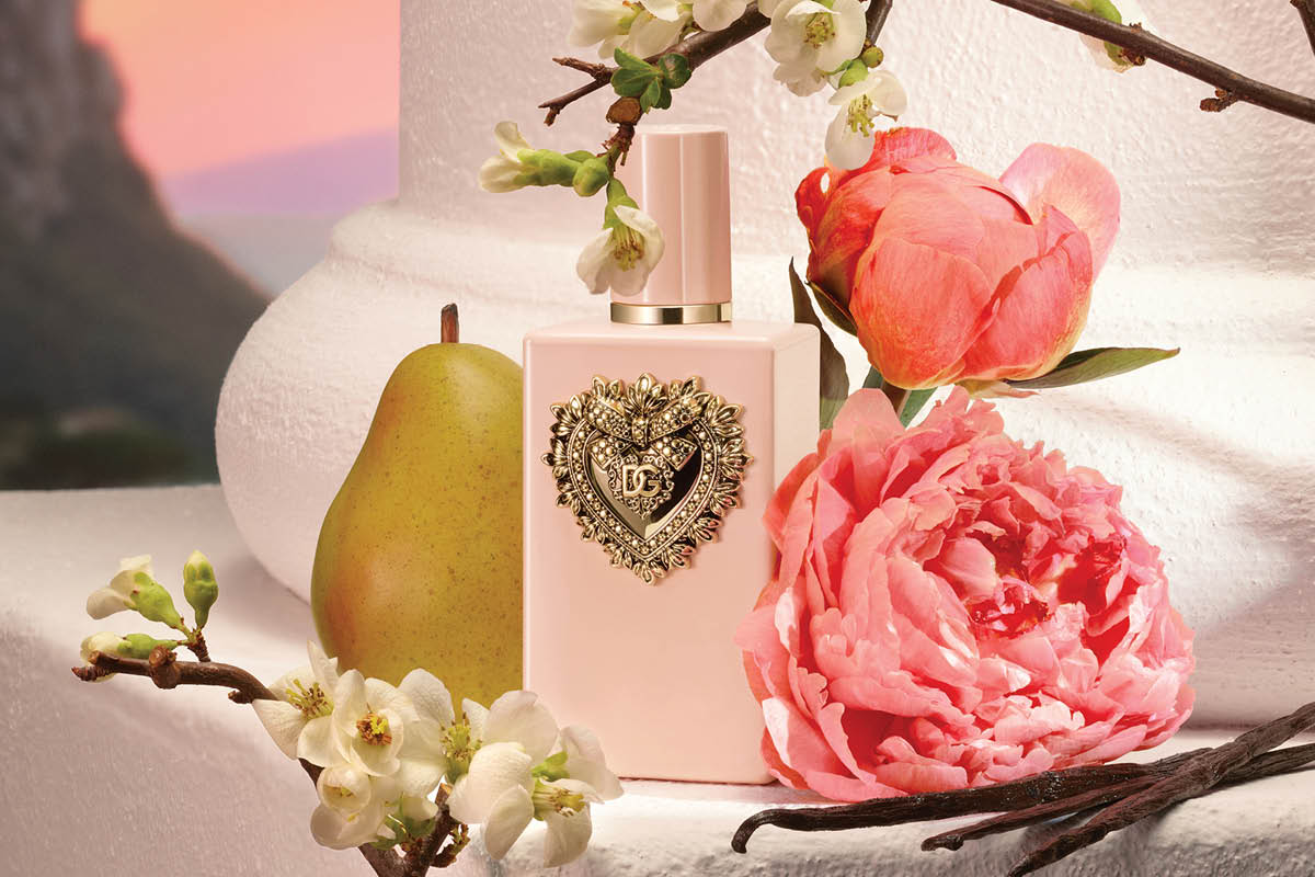 Dolce & Gabbana parfymeflaske i lys rosa med gulldetalj og hjerteformet DG-dekor er omgitt av blomster og frukt på en lys overflate.