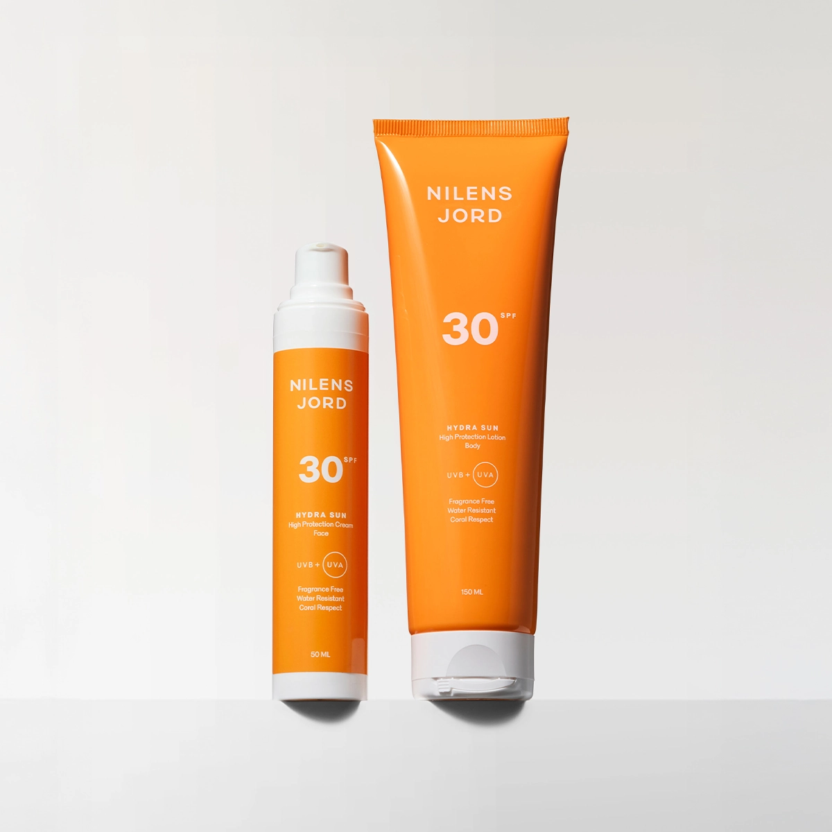 Kaksi oranssia Nilens Jord Hydra Sun SPF 30 -aurinkosuojatuotetta vaaleaa taustaa vasten. Pakkauksissa näkyvät tekstit “Nilens Jord”, “30 SPF”, “Hydra Sun”, “High Protection Cream”, “High Protection Lotion Body”, “UVB + UVA”, “Fragrance Free”, “Water Resistant”, “Coral Respect” sekä koot 50 ML ja 150 ML.