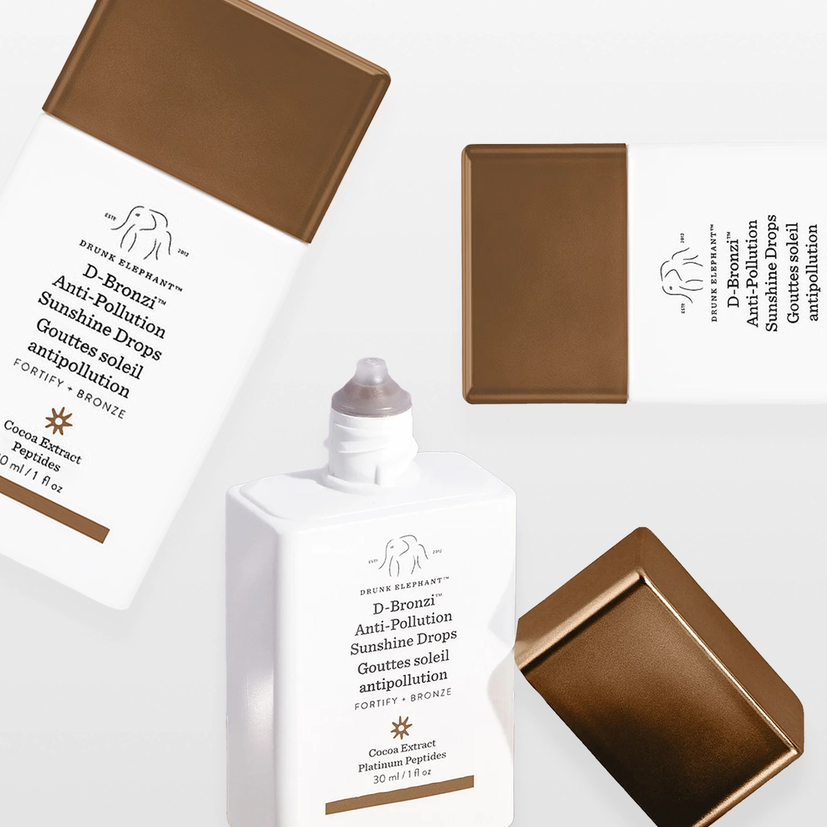 Tuotekuva, jossa useita Drunk Elephant D-Bronzi Anti-Pollution Sunshine Drops -pulloja ja korkkeja. Valkoisia, neliskulmaisia pulloja pronssinvärisillä korkeilla eri asennoissa vaaleaa taustaa vasten.