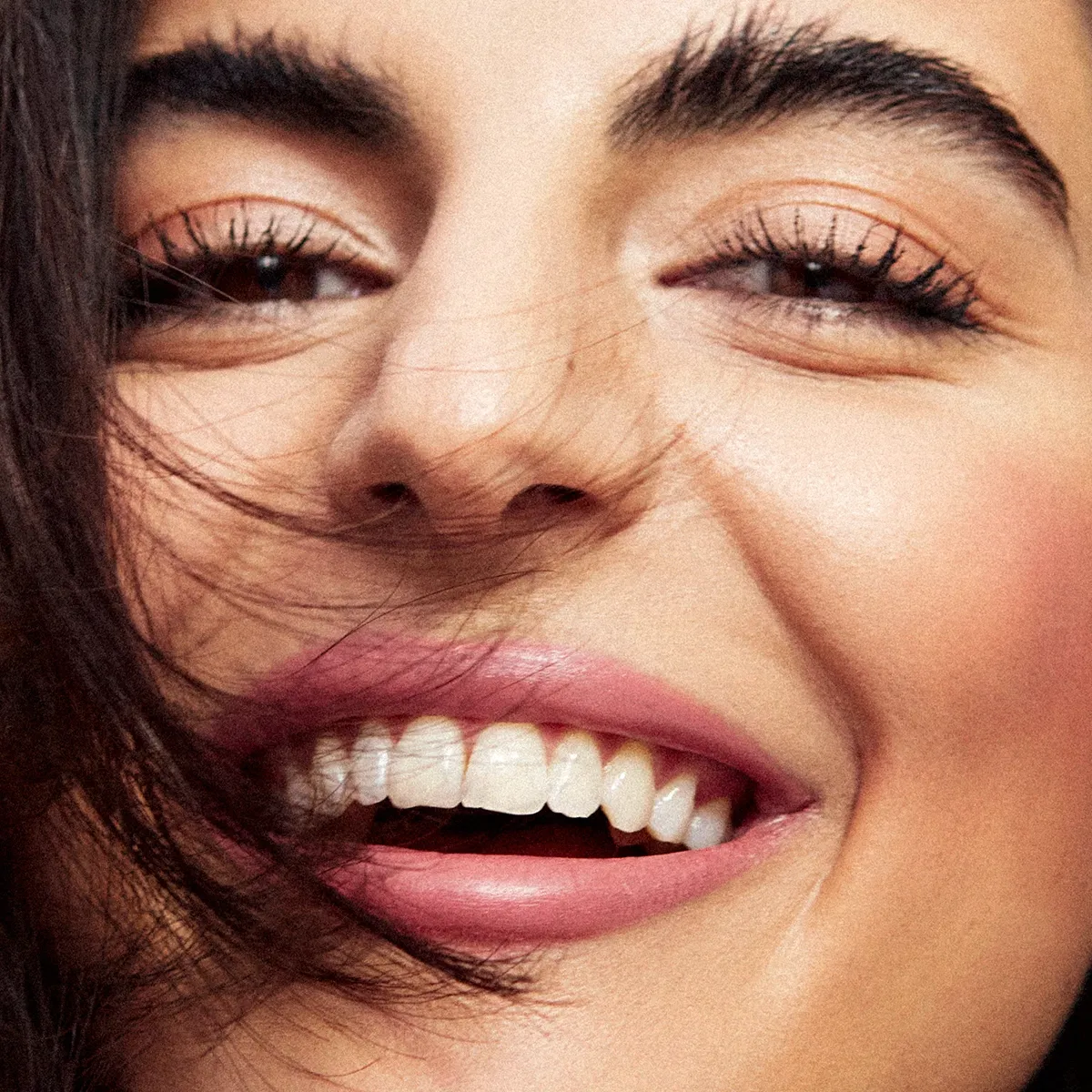 Närbild på leende kvinna med glansig hud, rosiga kinder och naturlig makeup mot rosa bakgrund.