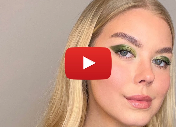 Nainen, jolla on vaaleat hiukset ja vihreä silmämeikki, hymyilee kameraan beigeä taustaa vasten.