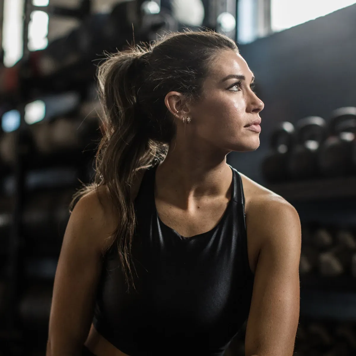 Kvinne i treningsklær sitter på treningssenter og ser til siden, med kettlebells i bakgrunnen.