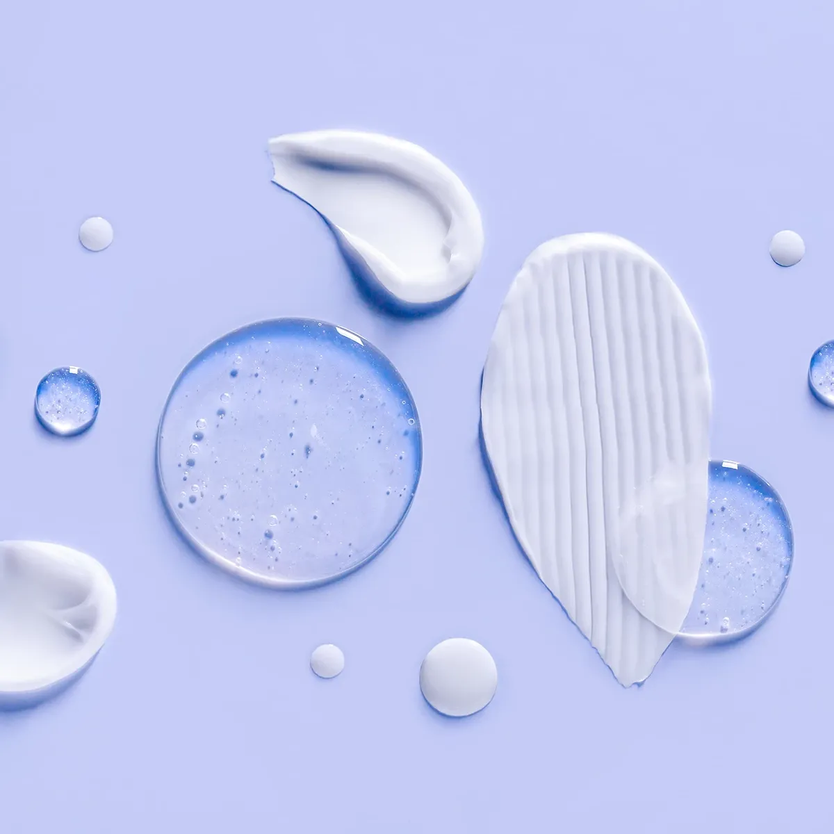 Ulike teksturer av hudpleieprodukter – kremer, geler og serum – spredt utover en lys lilla bakgrunn.