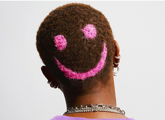 Baksiden av et hode med kort, mørkt hår der en rosa smileyform er laget i håret. Personen har flere øredobber og halskjeder mot en lys bakgrunn.
