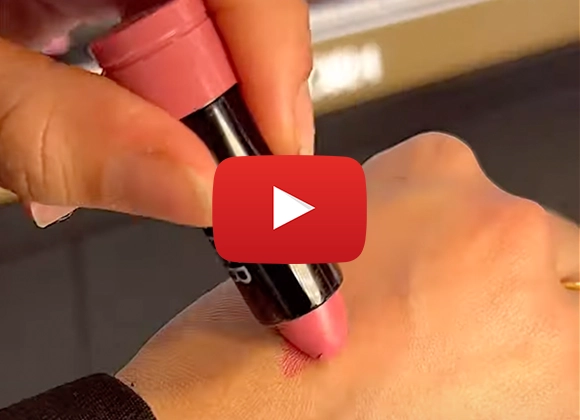 En hånd påfører en rosa leppestift på en annen hånd for å vise fargen. Et Youtube play-tegn er plassert midt på bildet.