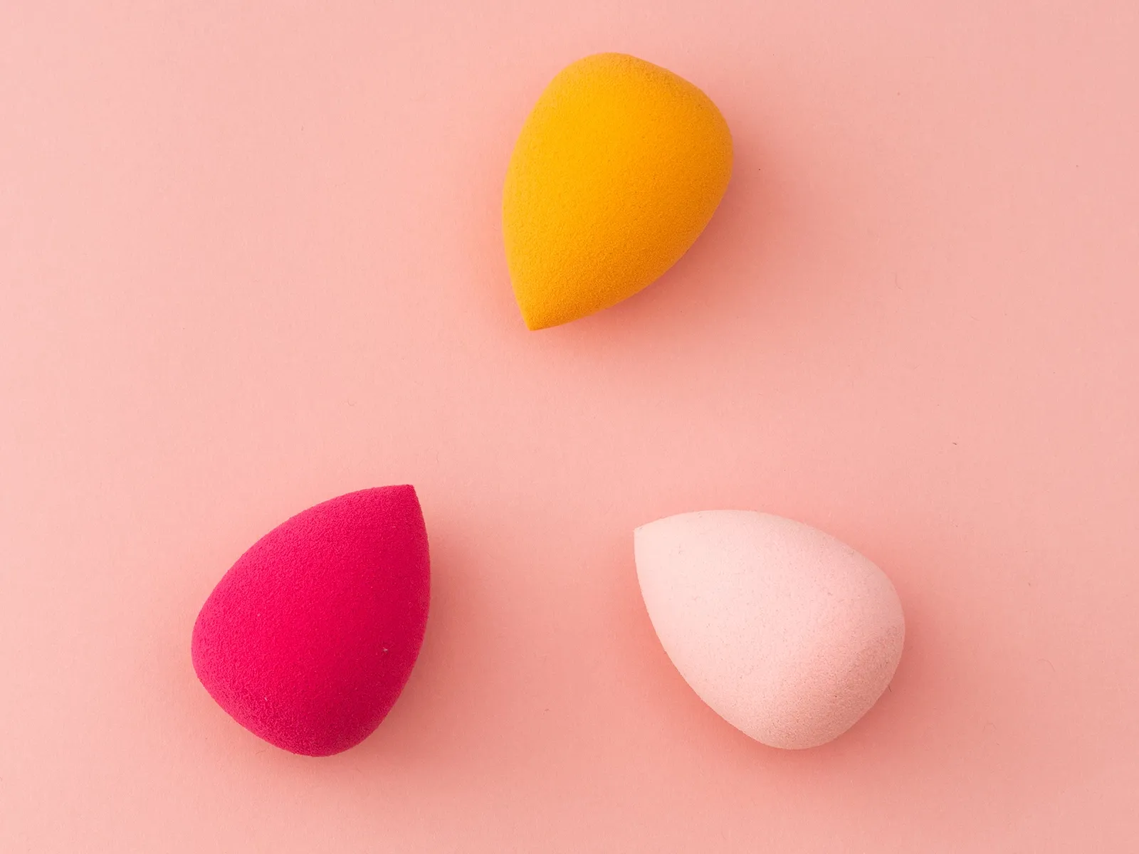 Tre sminksvamper i forskjellige farger (gul, rosa og rød) på en rosa bakgrunn.