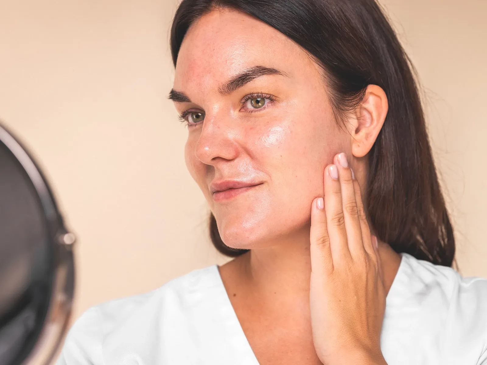 En kvinna med glansig hy och brunt hår masserar sin kind och speglar sig, mot en beige bakgrund.