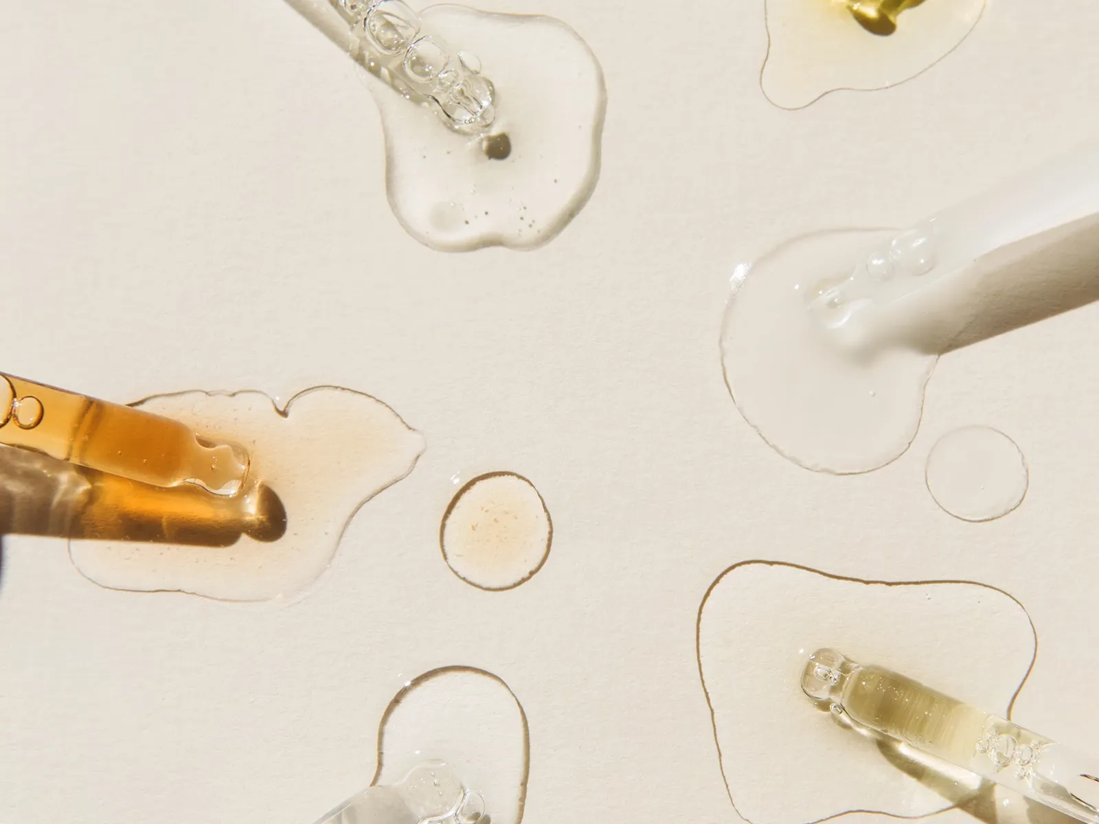 Droppar av sex olika vätskor på en beige texturerad yta i solljus