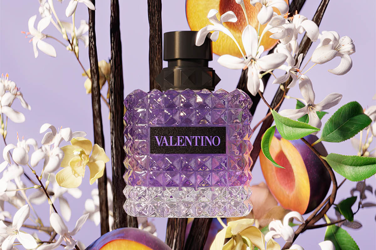 Lilla parfymeflaske med teksten “VALENTINO” plassert blant ferskener, grønne blader, hvite og gule blomster samt grener mot lilla bakgrunn. Flasken har fasettert design og mørk kork.