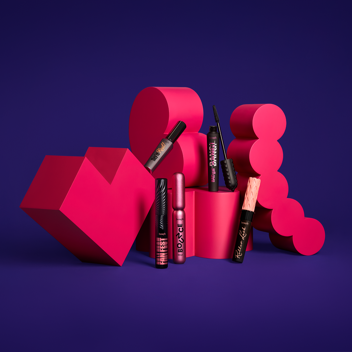 Fem forskjellige mascaraer fra Benefit Cosmetics står utstilt mot en fargerik bakgrunn med rosa og lilla geometriske former. Produktene har unikt og fargerikt design.