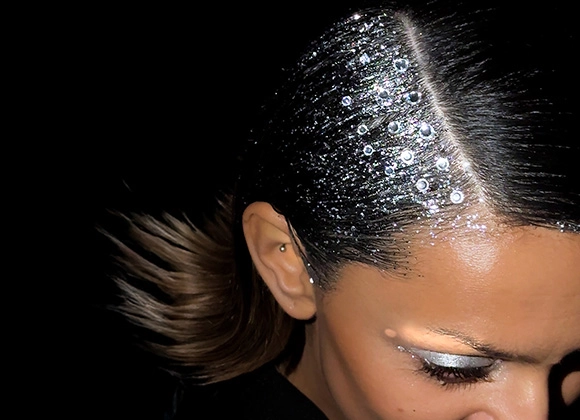 Lähikuva ylhäältä päin kuvattuna henkilöstä, jolla on taakse kiinnitetyt tummat hiukset. Hiusten jakausta pitkin on aseteltu pieniä kimaltelevia koristekiviä, jotka heijastavat valoa. Henkilöllä on hohtava luomiväri ja katse alaspäin tummaa taustaa vasten.