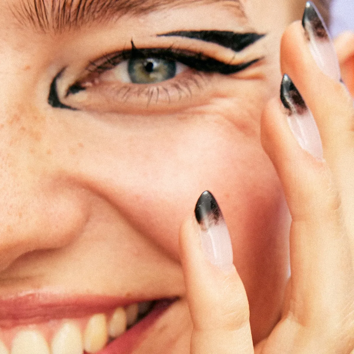 En person med svart ögonmakeup ler och håller handen vid ansiktet. Naglarna är målade med svart och vitt mönster.