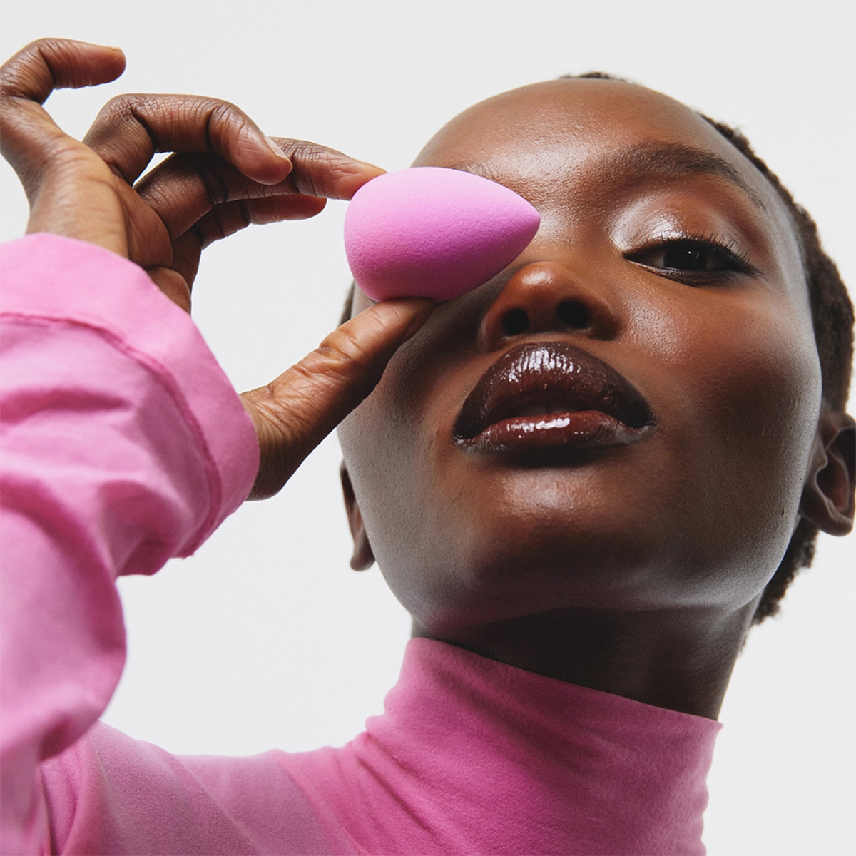 Närbild av en person i rosa polotröja mot ljus bakgrund. Personen håller en rosa sminksvamp framför det ena ögat med handen. Ansiktet är lätt vinklat uppåt och läpparna har blank makeup.