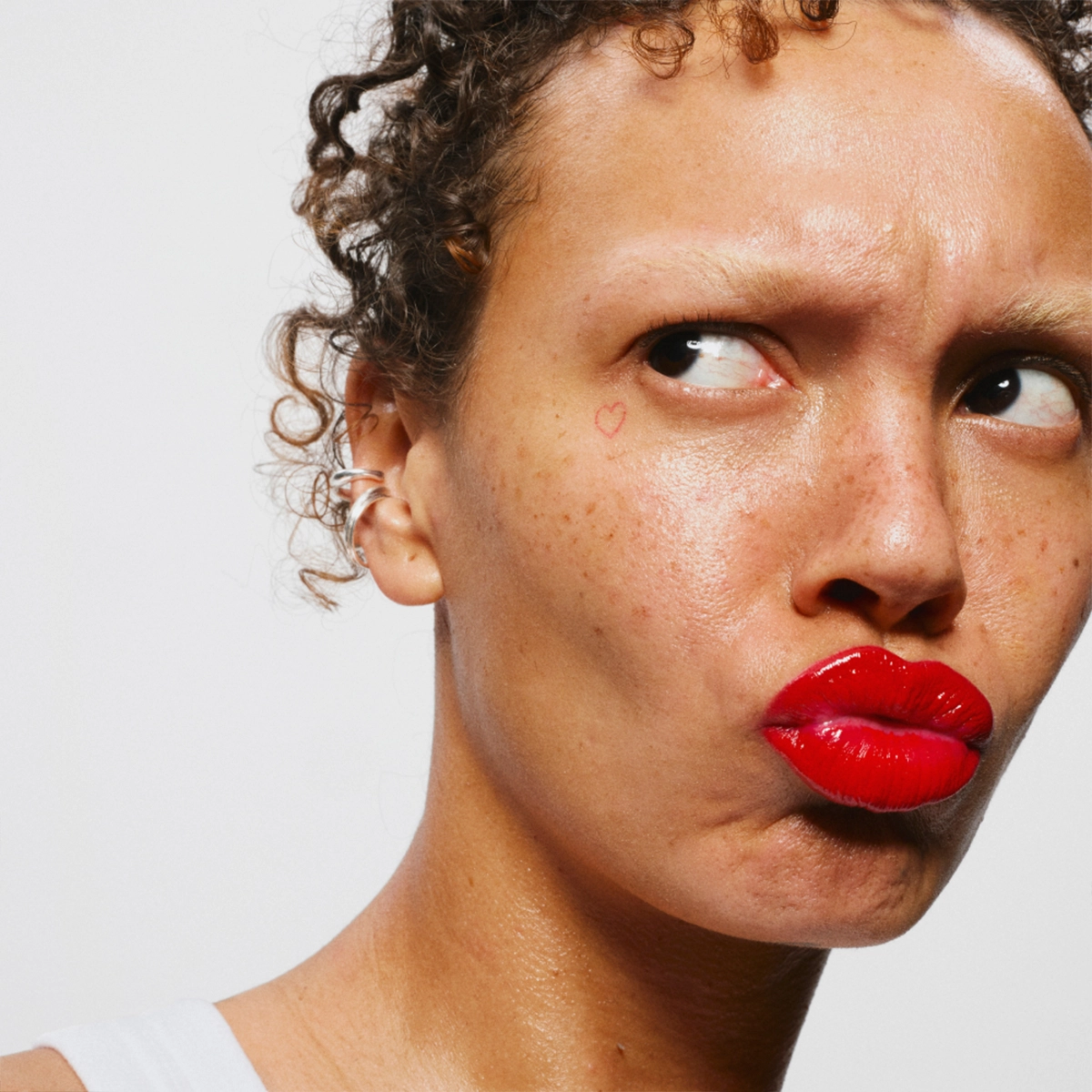 Person med röda läppar och naturlig makeup som tittar åt sidan med rynkad panna, med små fräknar och ett litet hjärta ritat på kinden mot ljus bakgrund.