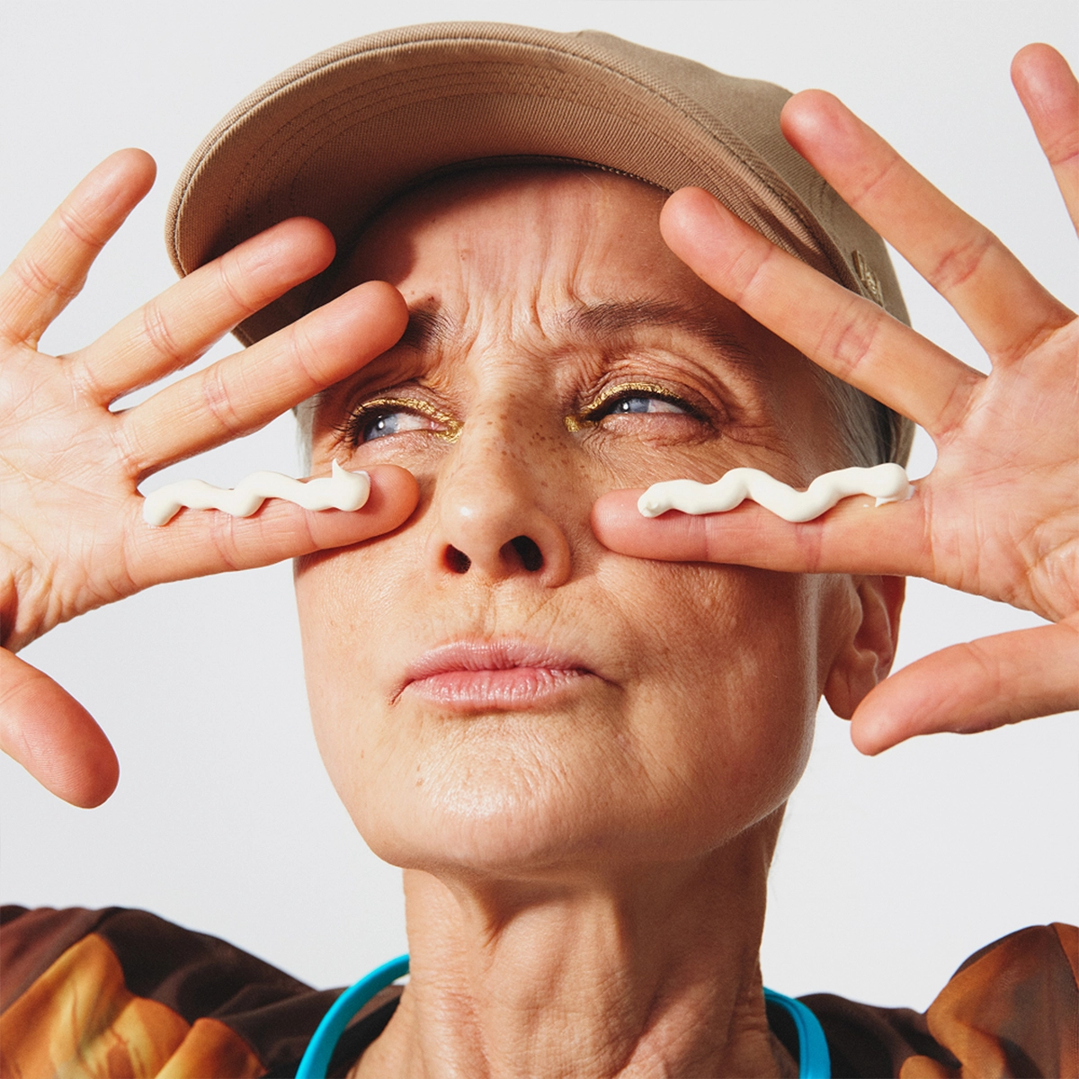 Person med beige keps applicerar två strängar ögonkräm under ögonen med fingertopparna. Närbild av ansikte med guldig ögonskugga och hudvård för ögonområdet mot ljus bakgrund.