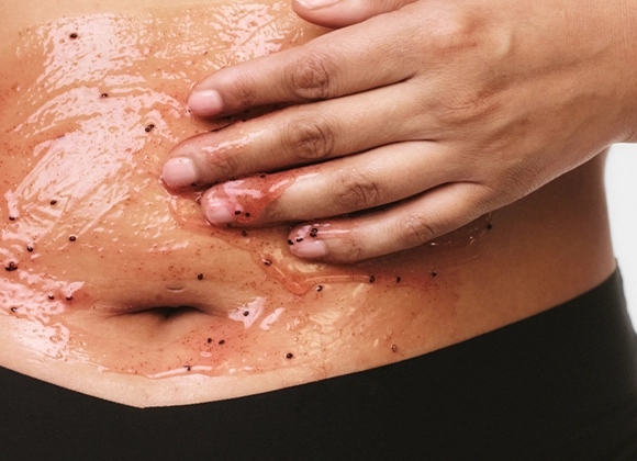 Närbild av en hand som applicerar kroppsskrubb med korn på magen, med glansig hud och synliga exfolierande partiklar inom hudvård.