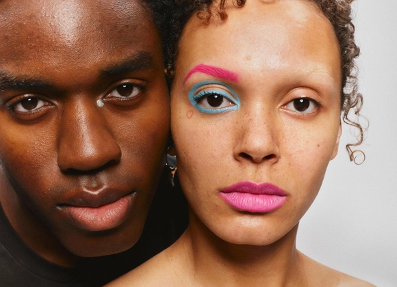 Två personer poserar nära varandra; en har färgstark och kreativ ögonmakeup i rosa och blått.