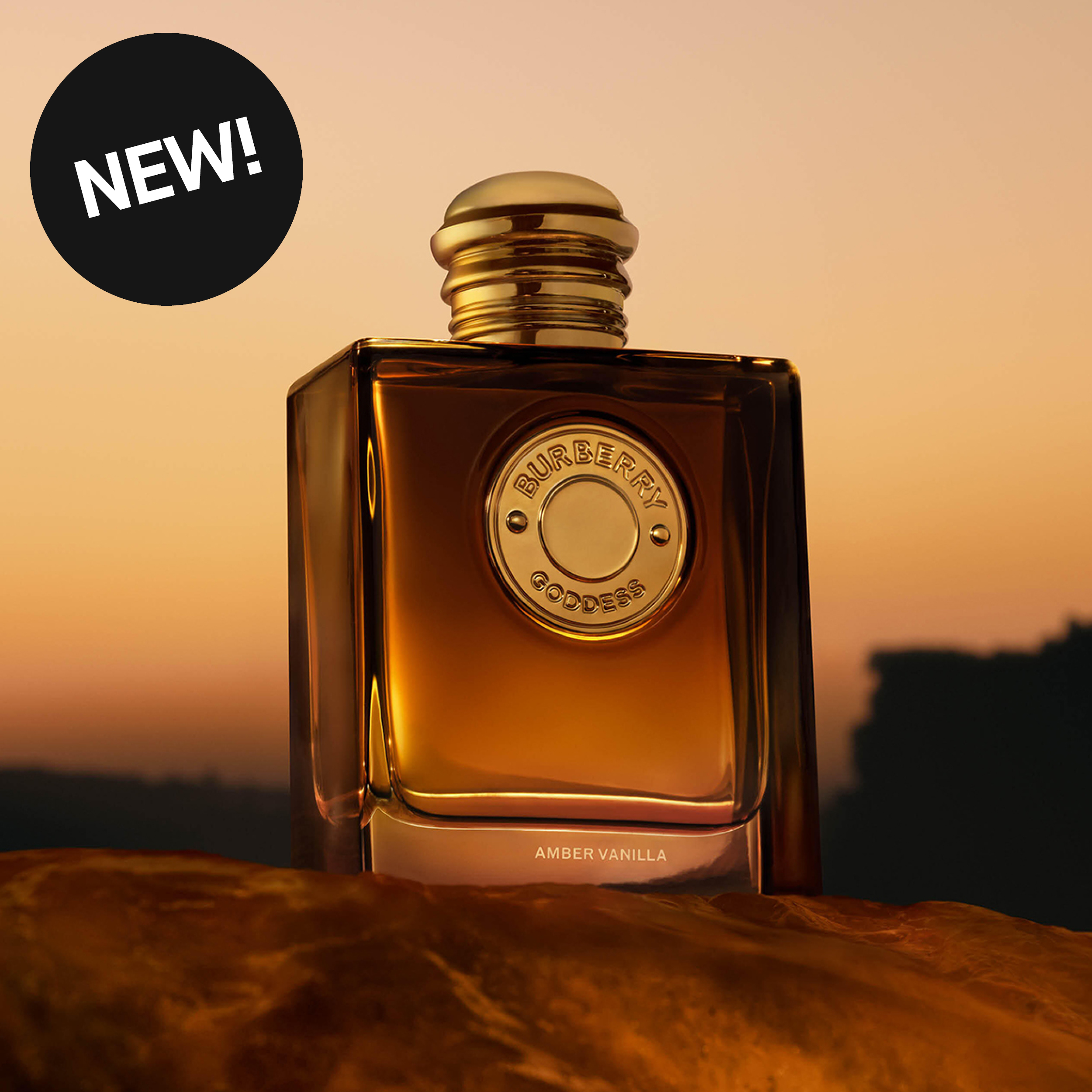 Burberry Goddess Amber Vanilla-parfyme i en ravfarget flaske med gullfarget kork står foran en varm solnedgang. Flasken har tekstene "BURBERRY", "GODDESS" og "AMBER VANILLA", og en svart sirkel med teksten "NEW!" vises øverst til venstre.