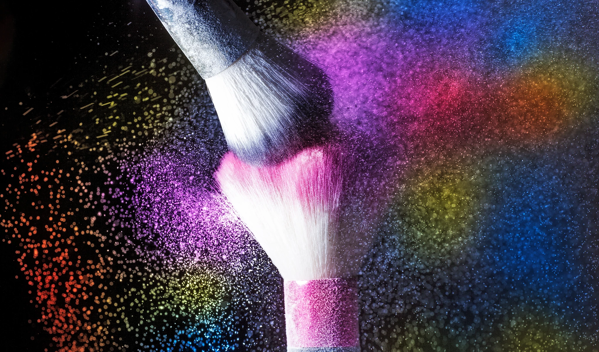 Två puderborstar mot svart bakgrund med massa färgglatt glitter i luften.