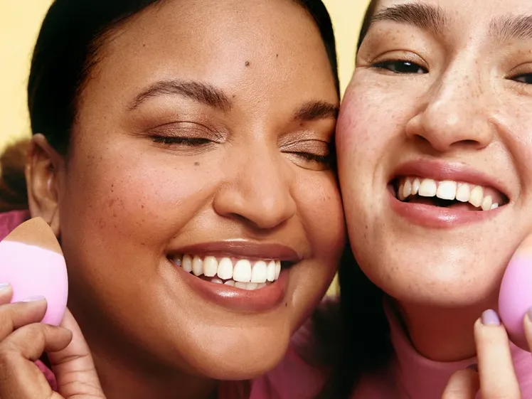 To kvinner står ved siden av hverandre, smilende og med sminkesvamper i ansiktet.