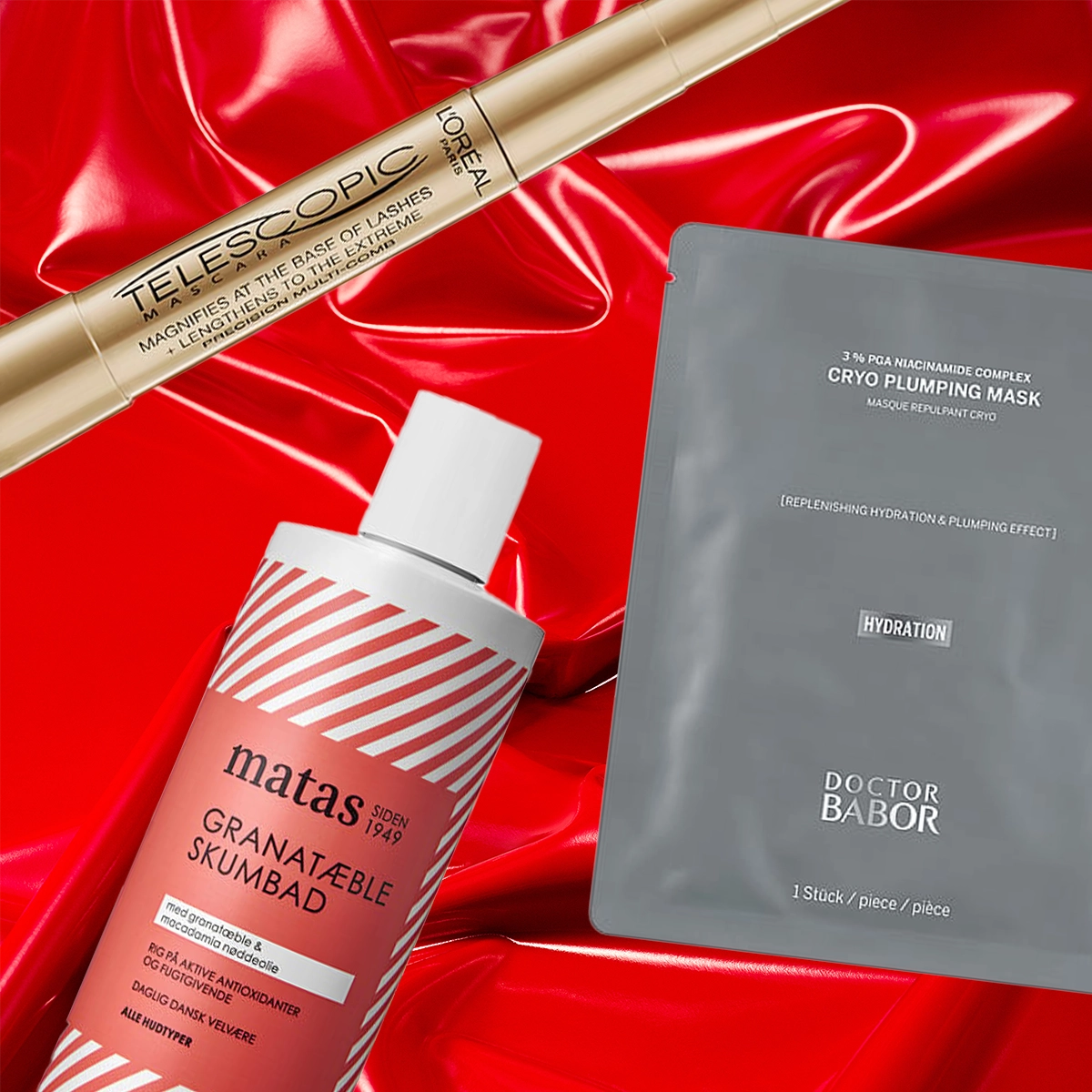 Tre skönhetsprodukter mot en röd, glansig bakgrund: en guldmaskara från L’Oréal Paris, en grå ansiktsmask från Doctor Babor och ett rött skumbad från Matas med granatäpple. Perfekta prisvärda julklappstips inom skönhet och hudvård.
